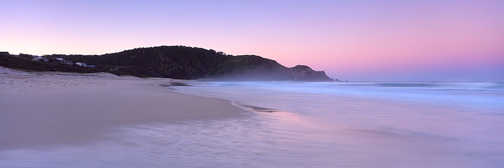 A serene beach scene is captured at dusk, showcasing soft pastel skies transitioning from pink to blue. Gentle waves roll onto the smooth shoreline, while a distant hill is framed by trees, creating a tranquil coastal view.