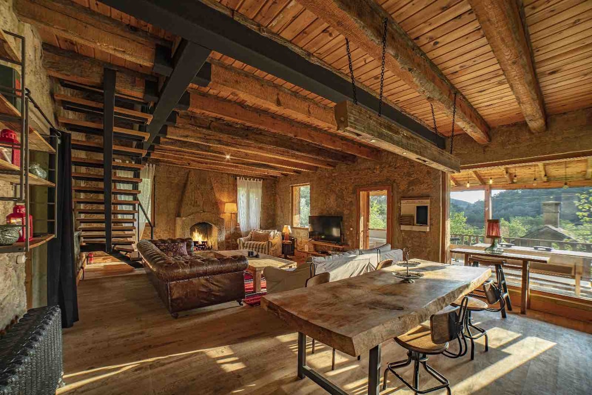 An open-concept living area features a spacious wooden dining table surrounded by chairs, with a comfortable sofa and armchairs arranged near a fireplace. Large windows allow natural light to fill the room, highlighting the wooden beams and rustic decor.