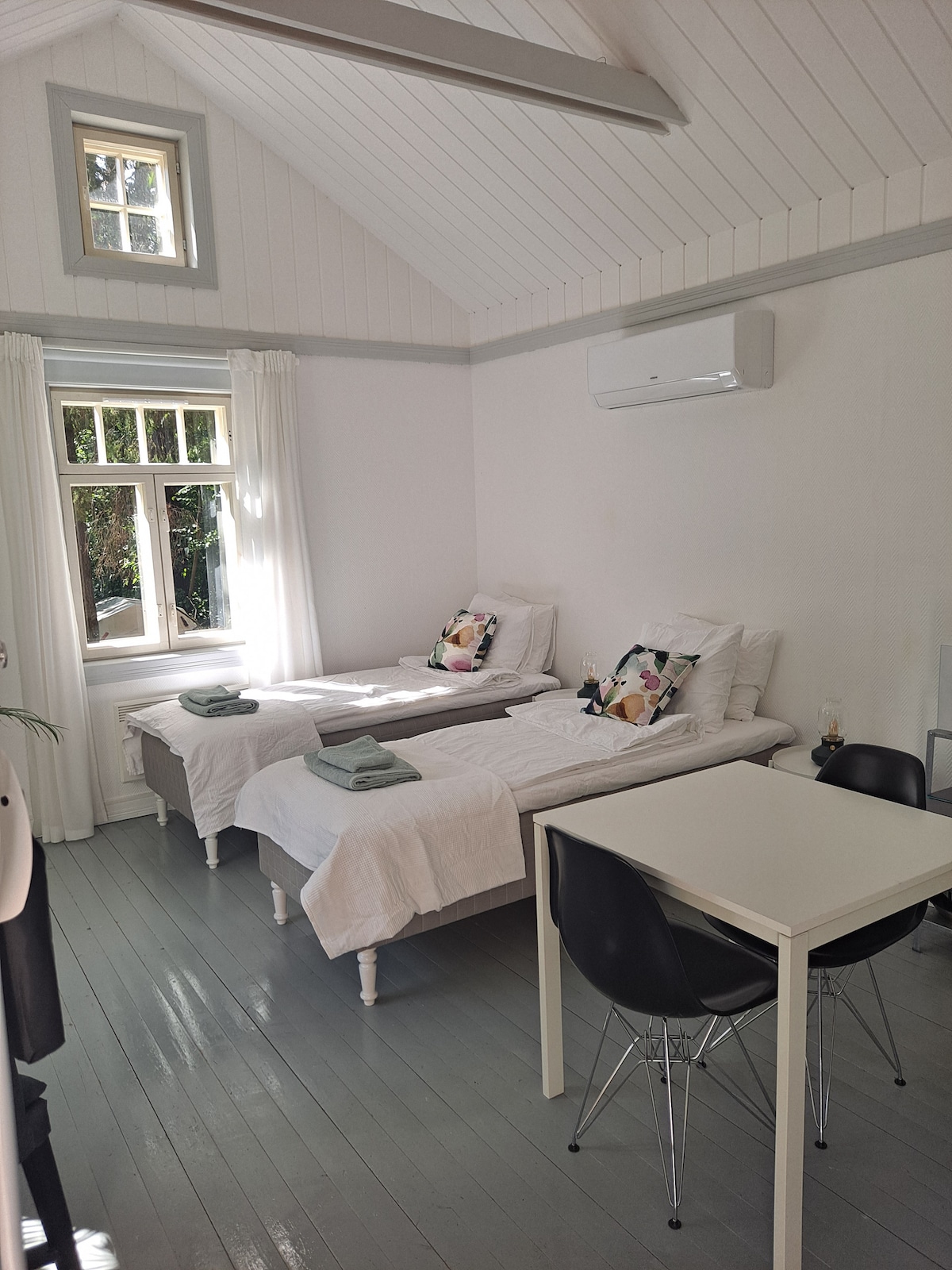 A bright and airy space features two single beds adorned with fresh linens and decorative pillows. A small dining table is positioned nearby, and natural light fills the room through a window with sheer white curtains. An air conditioning unit is mounted on the wall.