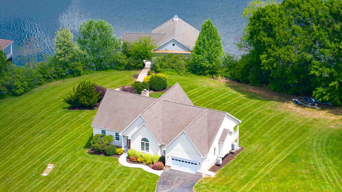 An aerial view of a lake resort home is captured, showcasing a well-manicured lawn and landscaping. The house features a spacious layout, with a driveway leading to a garage. Lake Anna reflects the surrounding greenery, enhancing the serene atmosphere of the property.