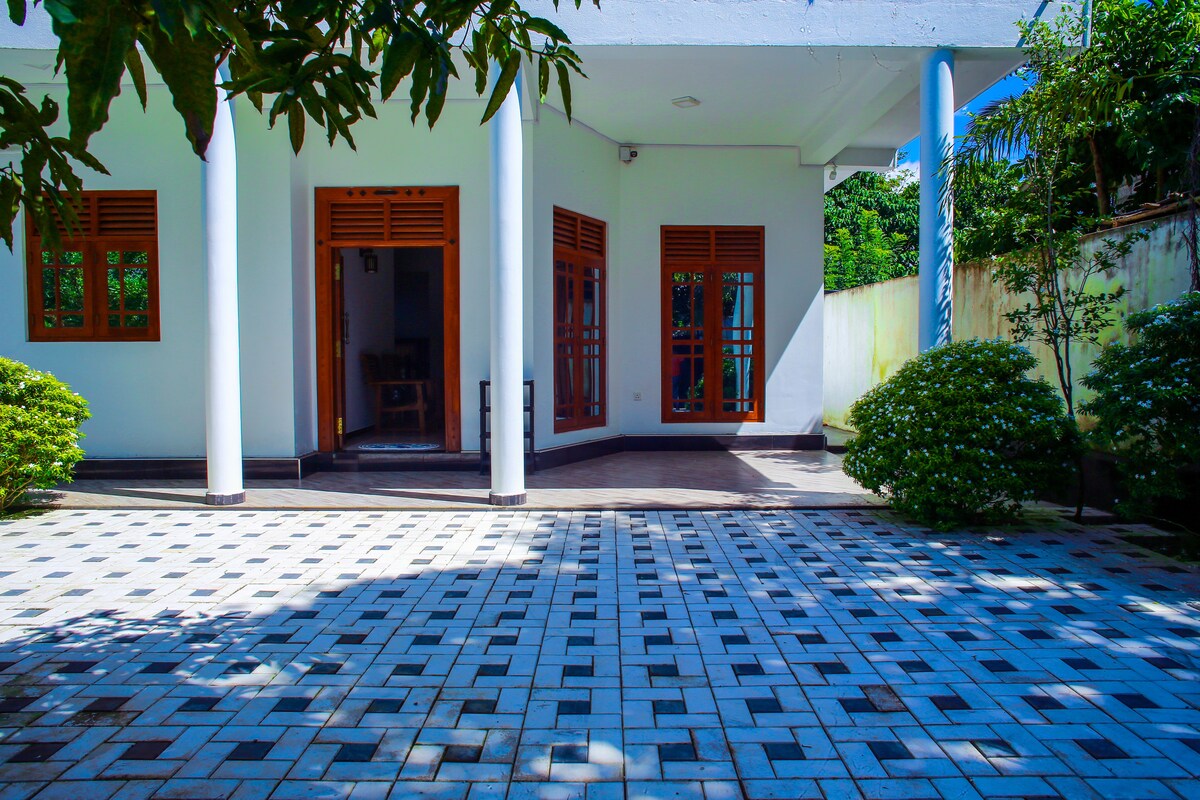 An open outdoor space is visible, featuring a tiled patio with a geometric pattern. Surrounding greenery, including well-maintained shrubs, frames the area. Large windows with wooden shutters provide natural light to the interior, enhancing the ambiance of the space.