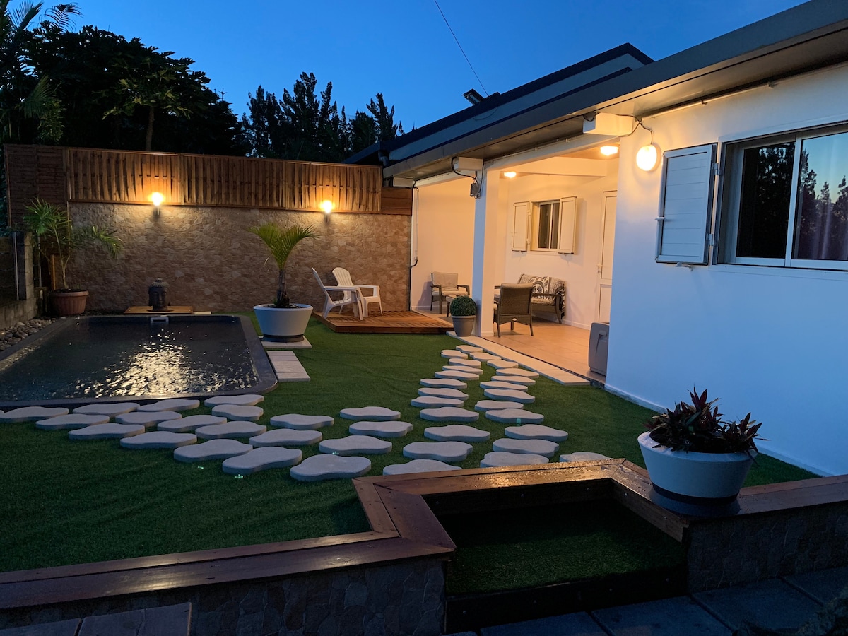 The outdoor space is illuminated by soft lighting, creating a serene atmosphere. A small pool is surrounded by decorative stones and artificial grass, while lounge chairs are positioned nearby for relaxation. Potted plants add a touch of greenery to the area.