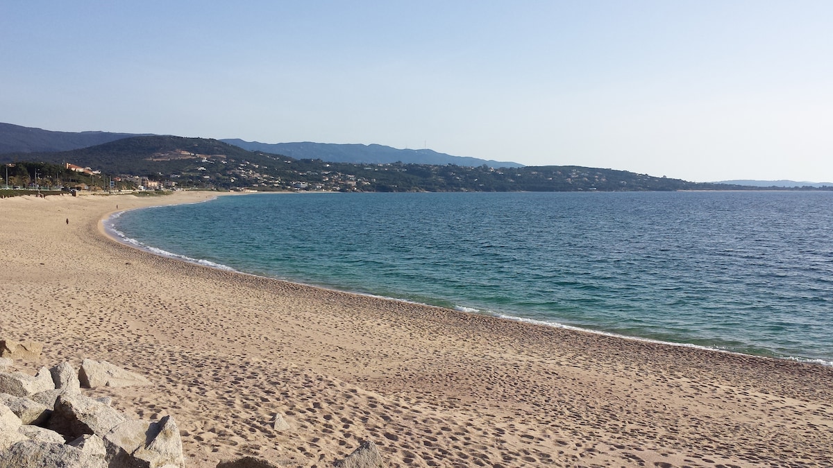 A wide sandy beach curves along the coast, meeting gently lapping waves. The shoreline is bordered by rocky formations, with hills visible in the background. The clear sky and calm water create a serene atmosphere, inviting individuals to explore the sandy landscape and enjoy the tranquil surroundings.