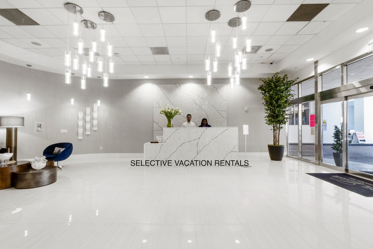 The reception area features a sleek marble front desk attended by two staff members. Bright pendant lights hang from the ceiling, illuminating the crisp white floors and walls. Lush greenery and modern seating are present, creating an open and welcoming environment.