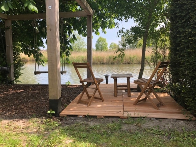 A serene outdoor seating area features two wooden chairs and a small table, positioned on a wooden deck surrounded by greenery. A swing hangs from a pergola, overlooking the water's edge adorned with tall reeds.