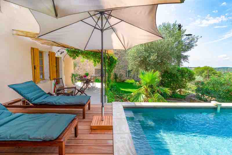 A shaded terrace area features two wooden lounge chairs with dark cushions, positioned beside a traditional salt-water pool. Lush greenery surrounds, including palm trees and olive trees, creating a serene atmosphere. The space invites relaxation and enjoyment of the tranquil outdoor environment.