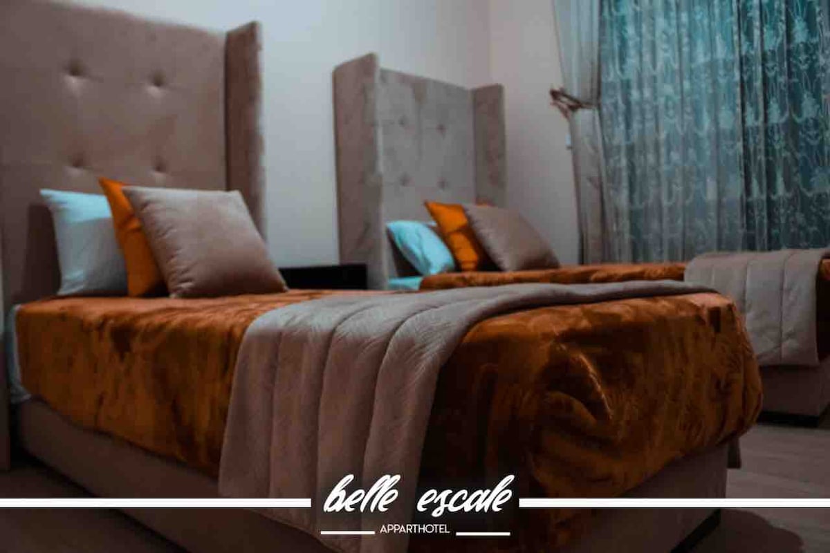 A well-appointed sleeping area is presented with two spacious beds, each adorned with decorative pillows and cozy blankets. Soft curtains frame the window, allowing natural light to filter in, creating a calm and serene ambiance in the room.