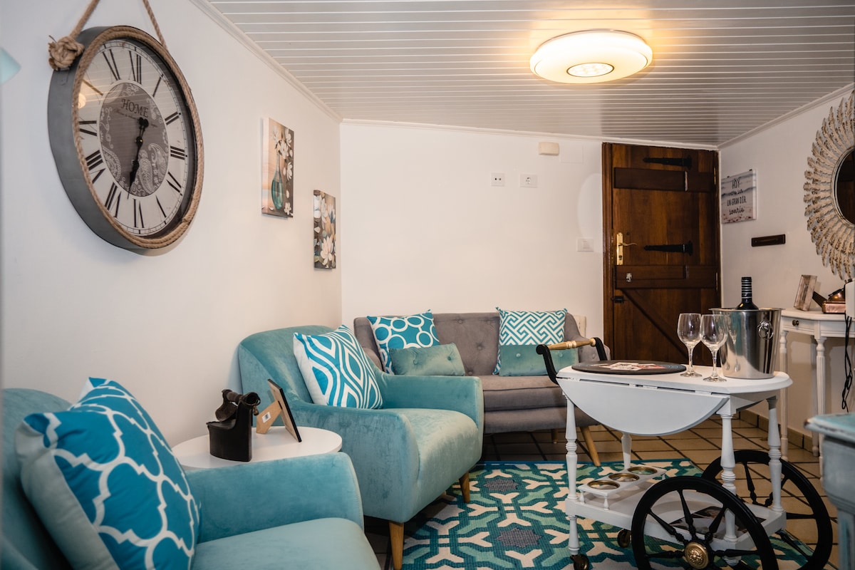 A inviting living area features a combination of contemporary and vintage styles, with framed artwork adorning the walls. Comfortable seating is arranged around a circular coffee table, complemented by decorative cushions. A large clock and a mirror add charm, while a detailed rug enhances the space.