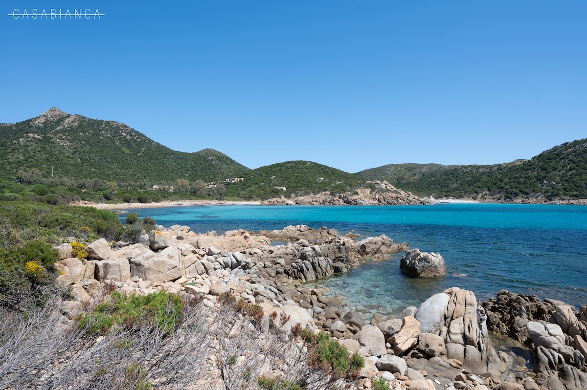 A coastal landscape features a rocky shoreline alongside clear turquoise waters. Green hills rise in the background, creating a natural, serene environment. The area showcases a blend of smooth stones and light vegetation, inviting exploration and enjoyment of the seaside scenery.