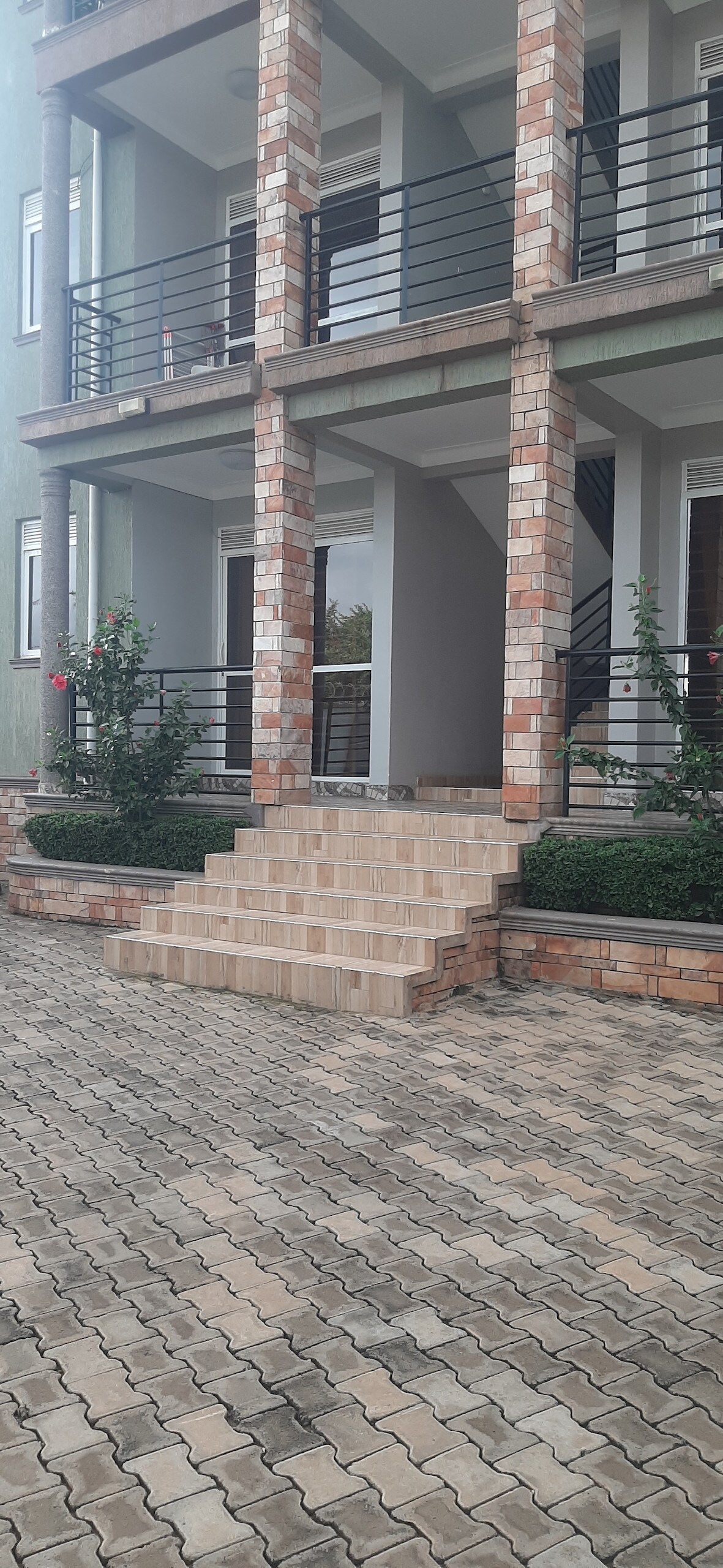 The exterior features a multi-story building with a brick facade and a set of steps leading up to the entrance. Lush greenery surrounds the base, providing a pleasant contrast to the stone-paved driveway.