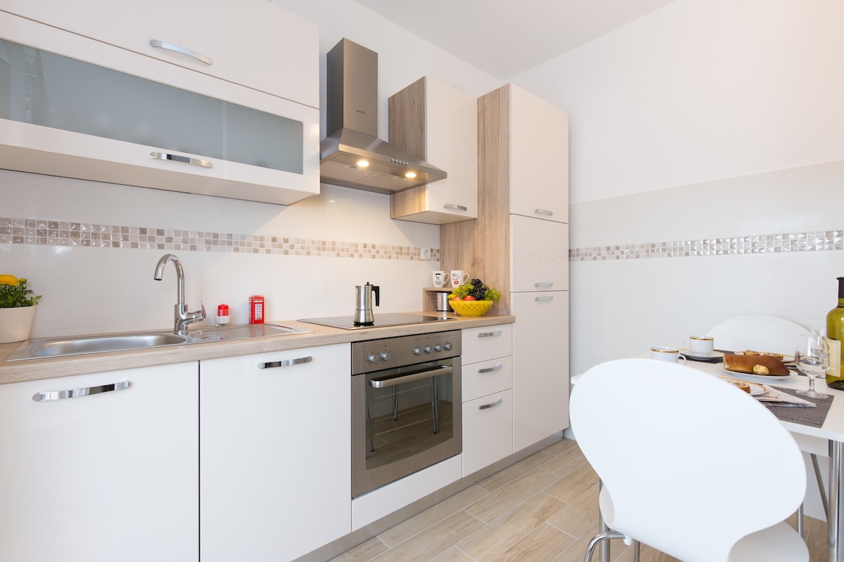 A modern kitchen is presented with sleek white cabinetry and a compact layout. Stainless steel appliances include an oven, stovetop, and kettle. A small dining table with white chairs is set for two, enhancing the functional and inviting atmosphere of the space.