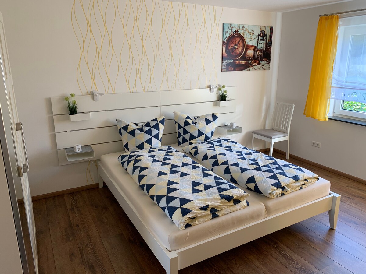 A well-lit bedroom is furnished with a spacious bed featuring geometric patterned bedding. Two bedside shelves hold small plants. A window with sheer yellow curtains allows natural light to enter, and a wooden chair is positioned nearby. Wall art enhances the decor.