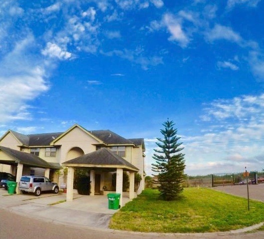 The exterior of a two-story home is depicted, featuring a well-maintained facade with a sloped roof. A tall tree stands in the front yard, and two vehicles are parked in the driveway. A clear blue sky stretches overhead, enhancing the setting.