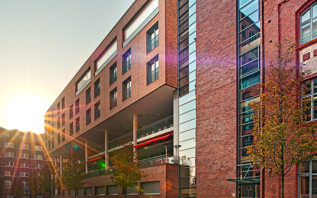 A modern building features a combination of brick and glass, reflecting the warmth of the sunset. Large windows offer glimpses of the indoor spaces, while trees are planted along the sidewalk, contributing to the inviting exterior environment.