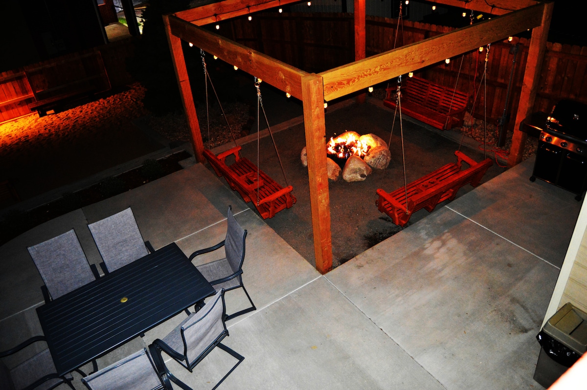 An outdoor gathering area is highlighted, featuring a wooden pergola with swings arranged around a central fire pit. A metal dining table with multiple chairs is placed nearby, creating an inviting space for socializing. String lights are visible, adding subtle illumination.