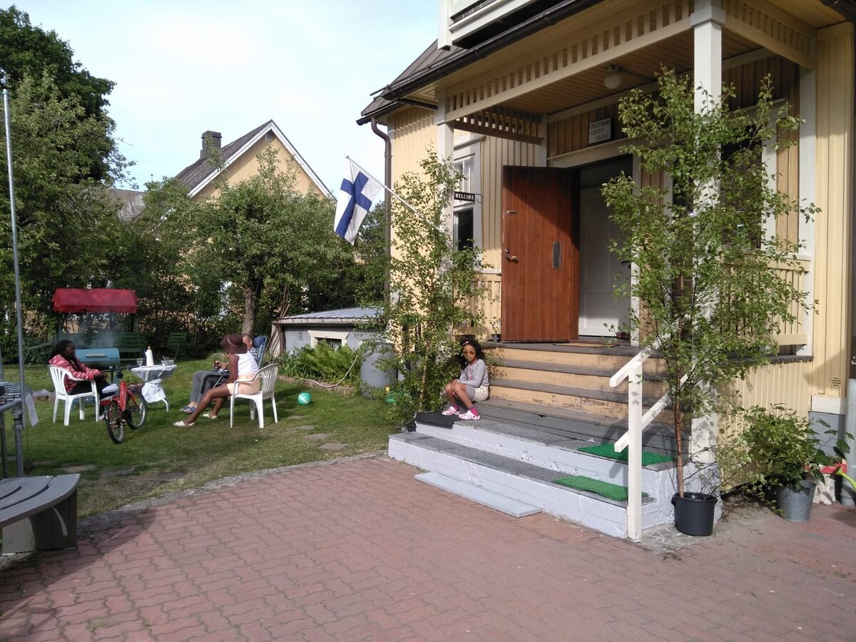 The entrance of a charming wooden house is shown, featuring a sturdy wooden door that opens onto a small porch. A seating area with white chairs is visible, surrounded by greenery. A Finnish flag billows gently in the breeze, adding a local touch.