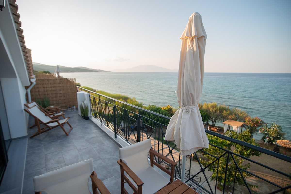 A sunlit balcony is furnished with wooden chairs and an upright umbrella, offering a view of the tranquil sea and distant hills. The soft colors of the landscape enhance the serene atmosphere, inviting relaxation near the water's edge.