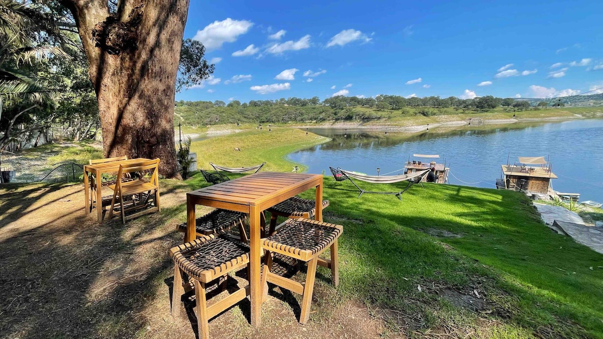 An outdoor area features a wooden table accompanied by four woven chairs, set against a lush green landscape. Hammocks are seen nearby, with the calm lake and distant tree line visible under a clear blue sky.