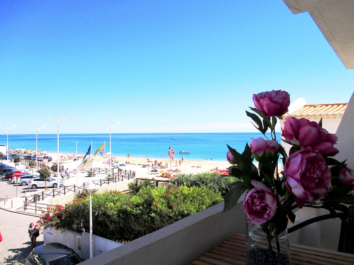 A scenic view from a balcony displays a sandy beach and clear blue waters, with people enjoying the sun. A vase of pink flowers sits on the wooden railing, adding a touch of color to the serene coastal landscape.