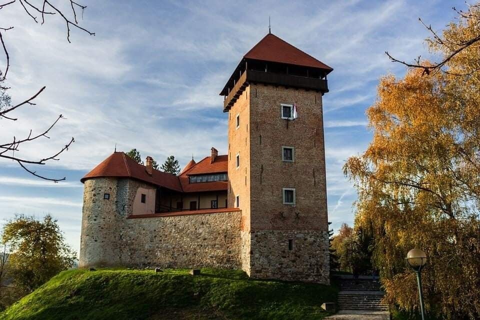 A historic stone tower stands prominently against a clear sky. Its sturdy walls and distinct red roof highlight its architectural features. Surrounding greenery and trees reflect autumn colors, enhancing the serene landscape, while winding pathways provide access to the structure.