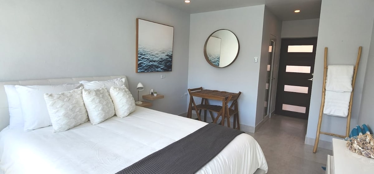 A well-lit bedroom features a queen-sized bed adorned with decorative pillows. A small wooden table is positioned nearby, while a round mirror reflects light. The room is complemented by soft gray walls and a minimalistic design, creating a calm and inviting space.