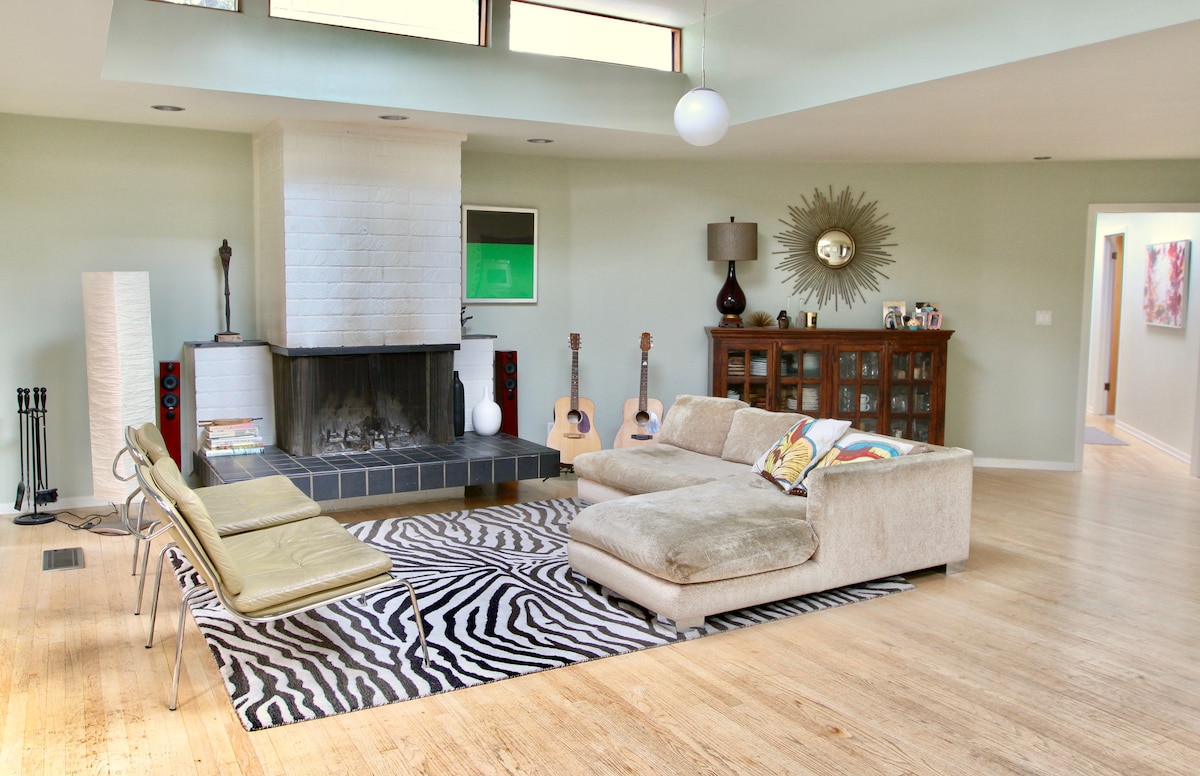 A spacious living area features large windows allowing natural light to fill the room. A modern sofa faces a brick fireplace, complemented by two sleek chairs and a decorative rug. Acoustic guitars and a sideboard with glass doors enhance the inviting ambiance of the space.