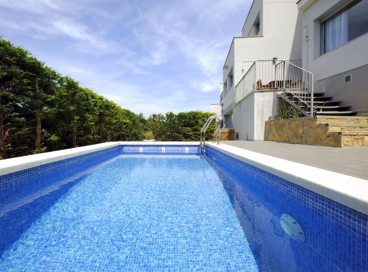 A private heated pool with clear blue water is surrounded by neatly trimmed greenery. Steps lead from the pool area to the house, which is partially visible in the background. A patio area can be seen adjacent to the pool.