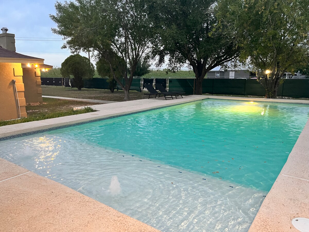 A large swimming pool is surrounded by a stone deck, featuring clear turquoise water that reflects the evening sky. Two lounge chairs are positioned along one side, and tall trees provide shade in the background, creating a peaceful outdoor setting.
