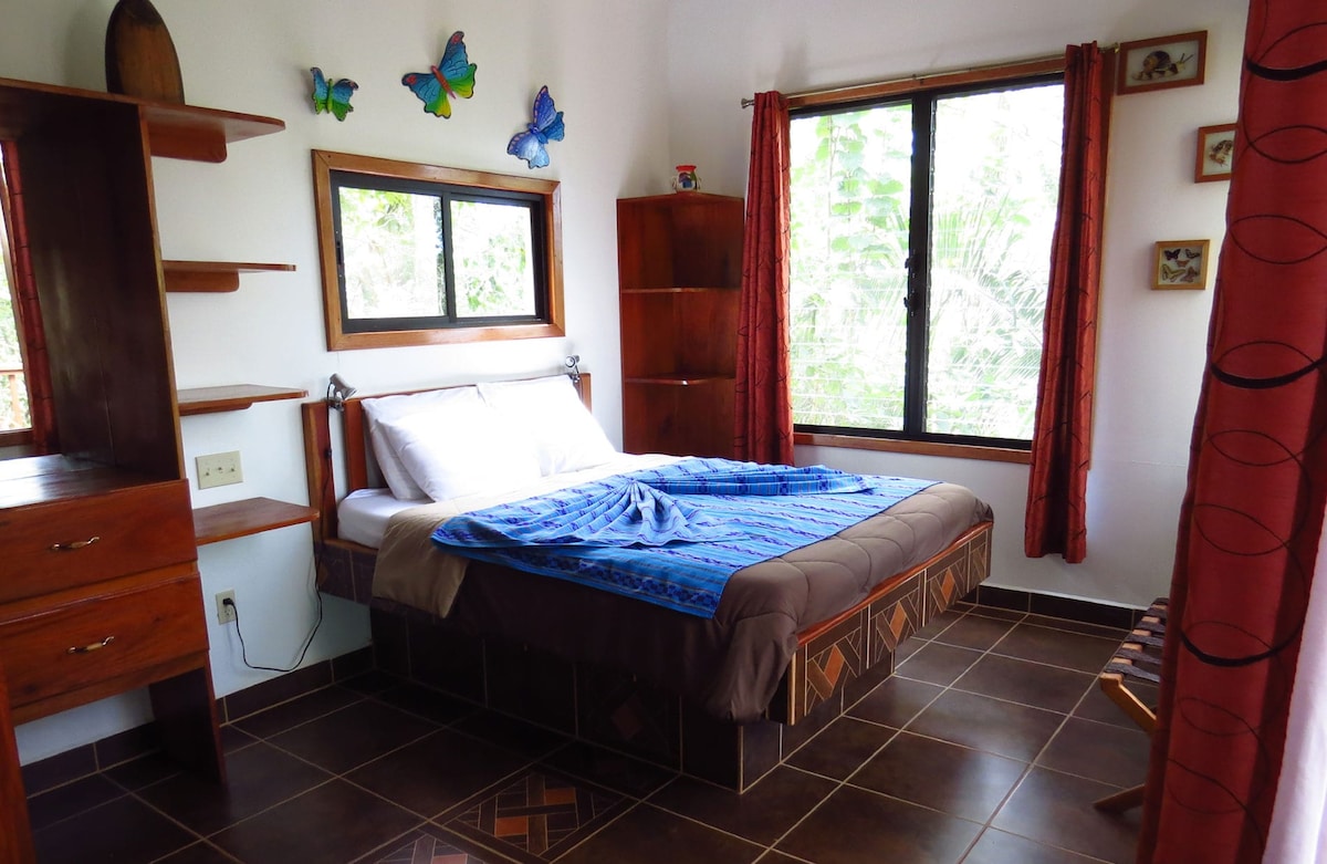A calming bedroom features a spacious bed adorned with a vibrant blue blanket. Natural light filters through large, framed windows, offering views of the surrounding greenery. Decorative butterflies are displayed on the wall, complementing the wooden shelving and furniture in the room.