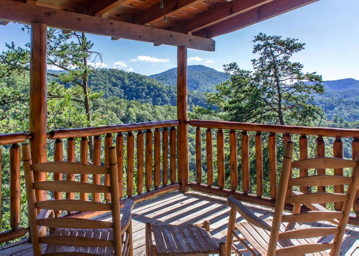 A wooden deck offers two rustic rocking chairs, positioned to face scenic mountain views. Lush greenery and rolling hills extend into the distance, framed by trees. The warm tones of the wood structure complement the natural setting.