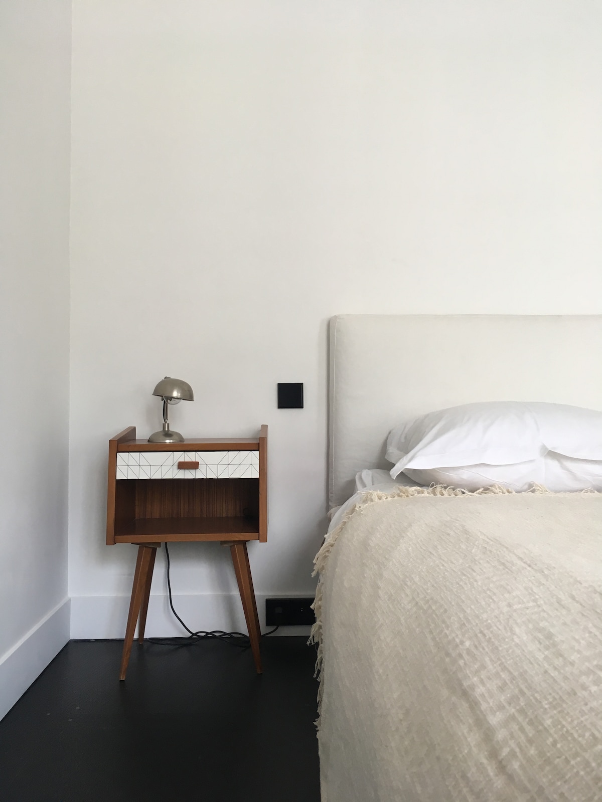 A minimalist bedroom corner is depicted, featuring a spacious bed with white bedding. A wooden nightstand with a geometric design and a small, metallic lamp is positioned beside the bed. The room has a clean, white wall and smooth, dark flooring.