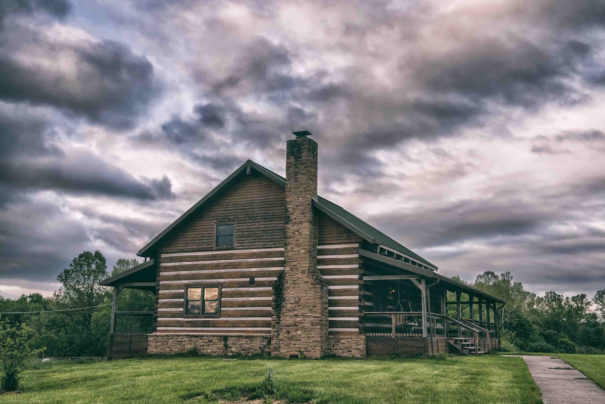 The exterior of a rustic cabin is depicted, featuring a combination of log and stone construction. A prominent chimney rises from the side, and a covered porch extends across the front, with large windows reflecting the overcast sky.