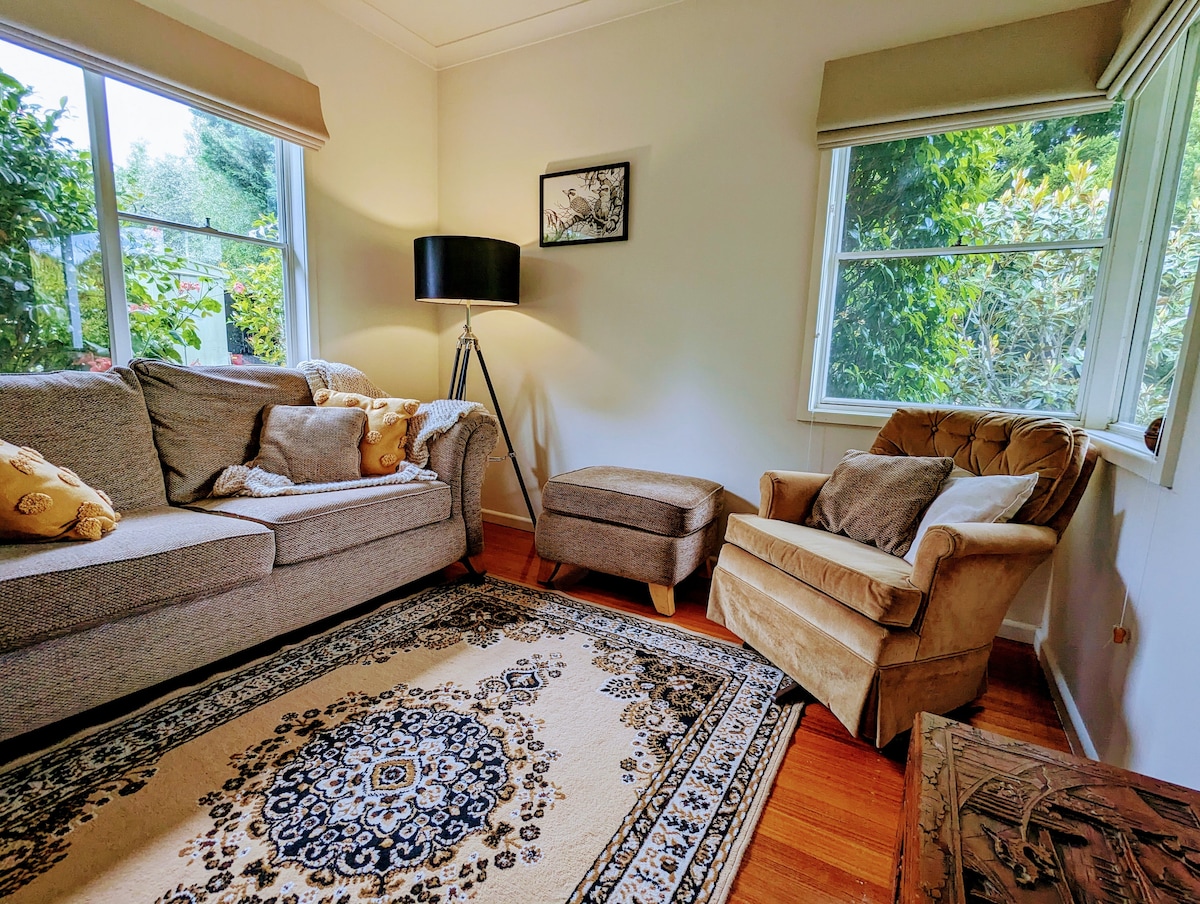 A cozy lounge area features a grey sofa adorned with decorative cushions, paired with a plush armchair. Large windows allow natural light to brighten the space, while a patterned area rug adds warmth underfoot. A lamp stands next to the sofa, providing additional lighting.