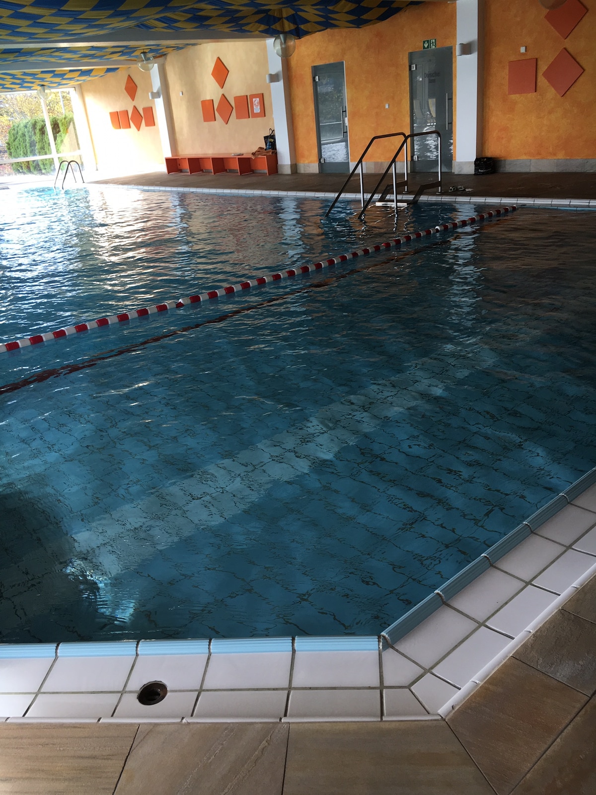 An indoor swimming pool is visible, featuring clear blue water reflecting overhead light. The pool area is accented by a colorful patterned ceiling and orange walls, with several steps leading into the water. Seating is available along the edges of the pool.