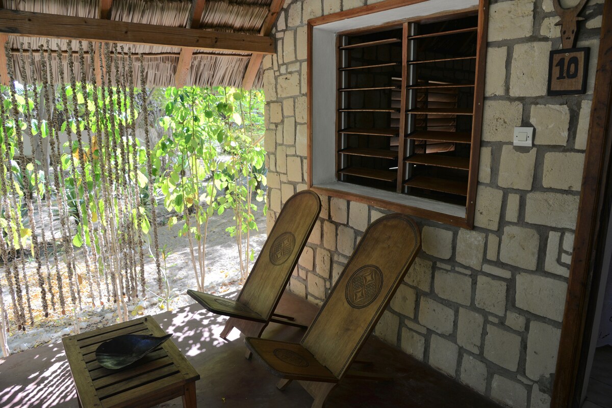 A small outdoor seating area features two wooden chairs with carving details, accompanied by a simple wooden table. Natural light filters through green foliage, highlighting the stone wall and creating a relaxed ambiance just outside the entrance.