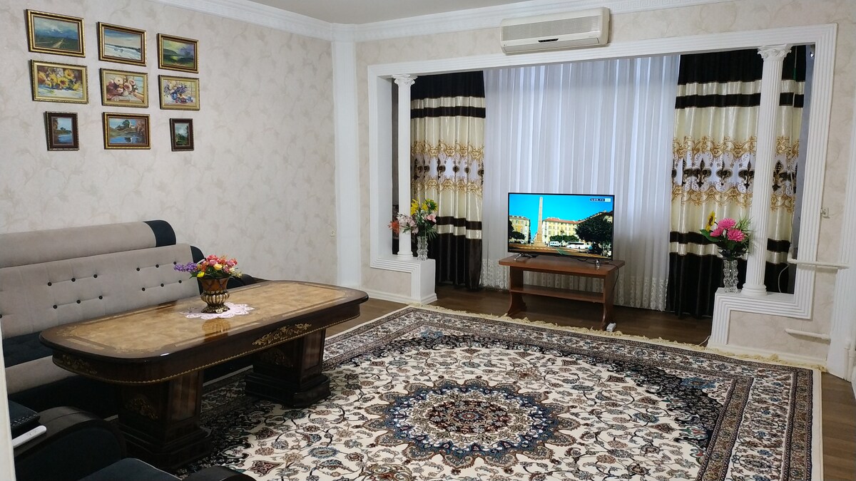 The living room features a large, patterned area rug centered under a wooden coffee table. A comfortable sectional sofa is positioned nearby. A smart TV is mounted on a wooden stand, complemented by floral arrangements and framed artwork on the walls. Natural light filters in through sheer curtains.
