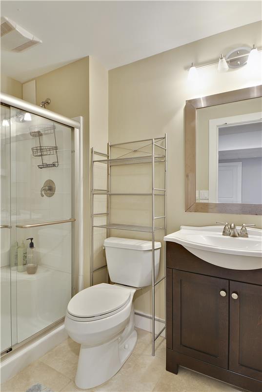 The bathroom features a glass-enclosed shower, a modern toilet, and a dark wood vanity. A sleek mirror is mounted above the sink, and a metallic shelving unit offers additional storage. Soft lighting highlights the neutral wall tones and tiled flooring.