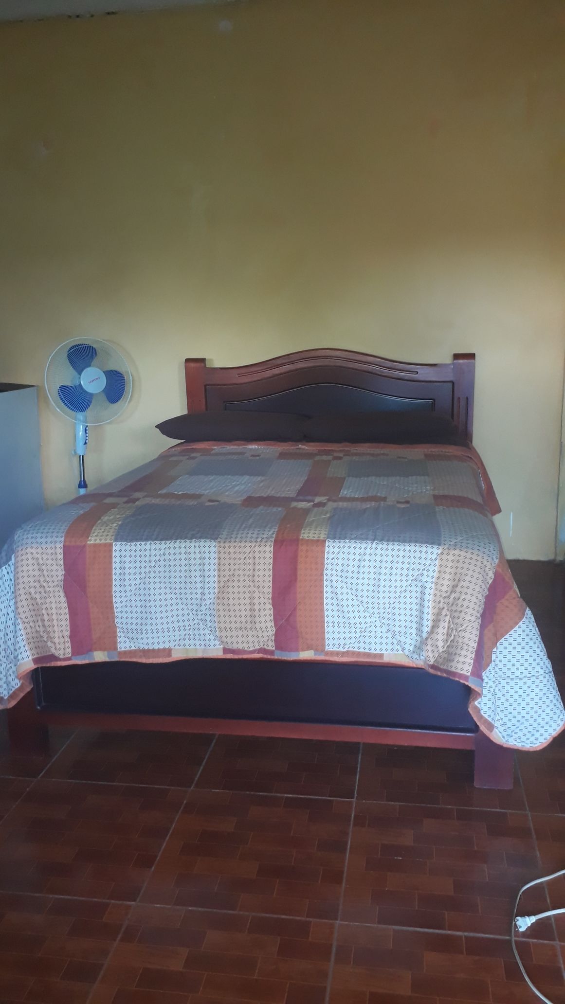 A comfortable sleeping area is highlighted by a wooden bed frame adorned with a patterned quilt. A standing fan is positioned nearby for added comfort, and the walls feature a soft yellow hue, creating a serene atmosphere. The tiled floor reflects a warm, inviting tone.