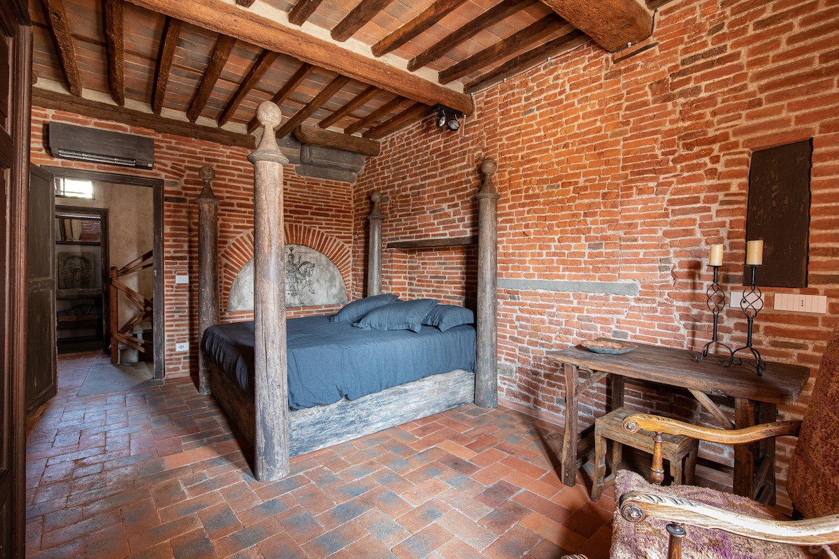 A spacious bedroom is characterized by exposed brick walls and wooden beams overhead. A large bed with a dark duvet is positioned centrally, accompanied by a rustic wooden table and an antique chair. Soft, natural light filters in, enhancing the authentic medieval ambiance.