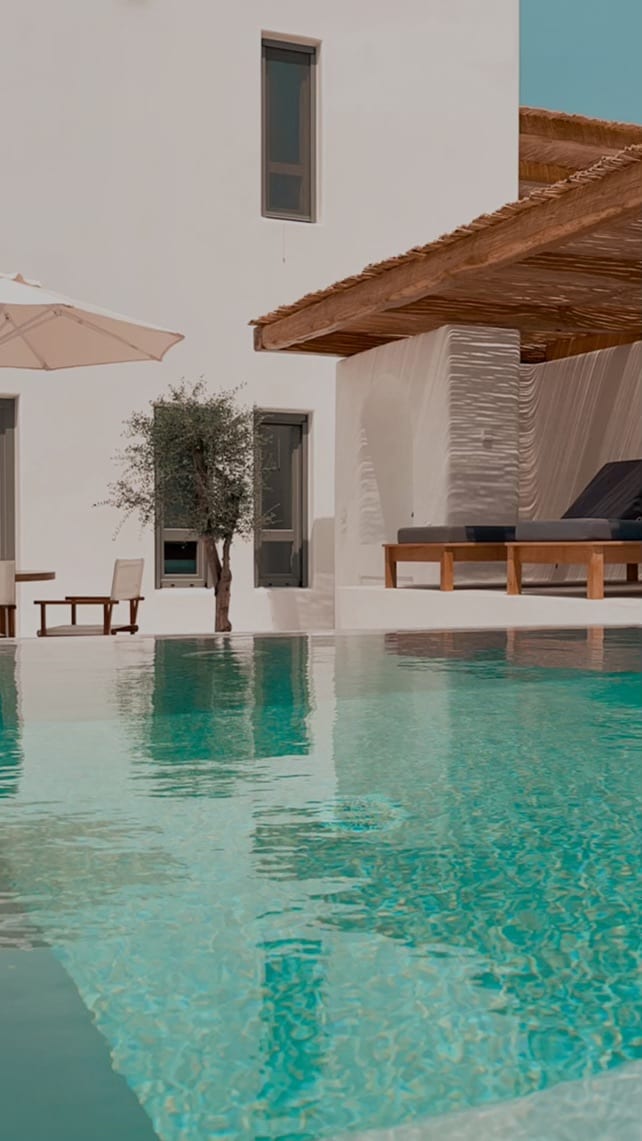 A private pool is displayed with clear blue water reflecting the building's exterior. A shaded area with wooden seating is visible nearby, alongside a small olive tree and an umbrella. The minimal architecture highlights clean lines and a serene outdoor environment.