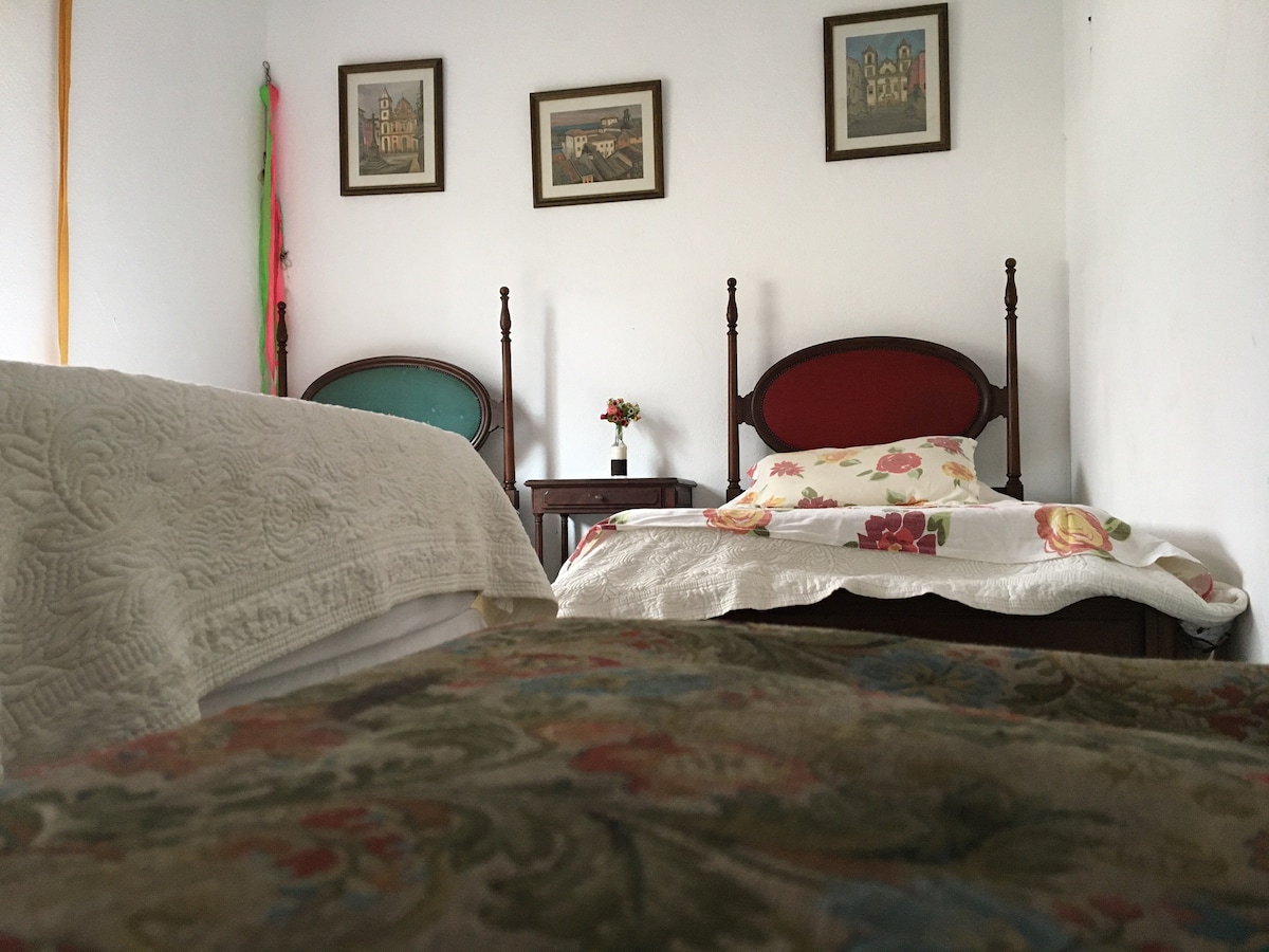 A cozy sleeping area features three single beds, adorned with floral-patterned bedding. A small bedside table holds a vase with fresh flowers. The walls are decorated with framed artwork, adding a touch of character to the space.