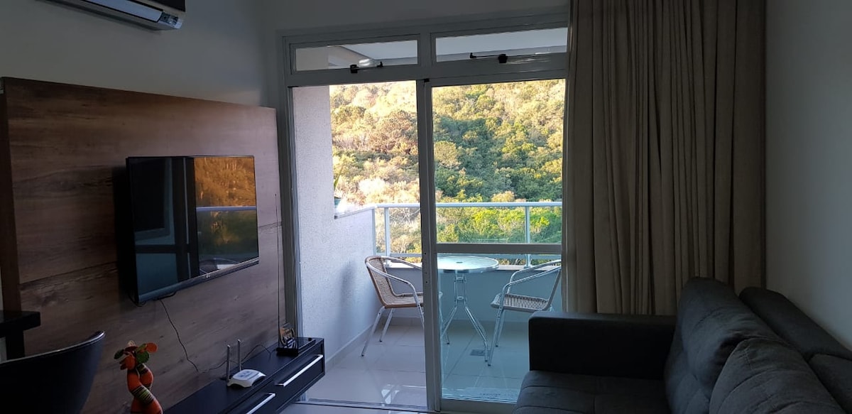 A living area is visible, featuring a sleek television mounted on a wooden panel. A comfortable gray sofa faces the TV. Large glass doors lead to a balcony with a scenic view of greenery. Natural light floods the space, enhancing its openness.