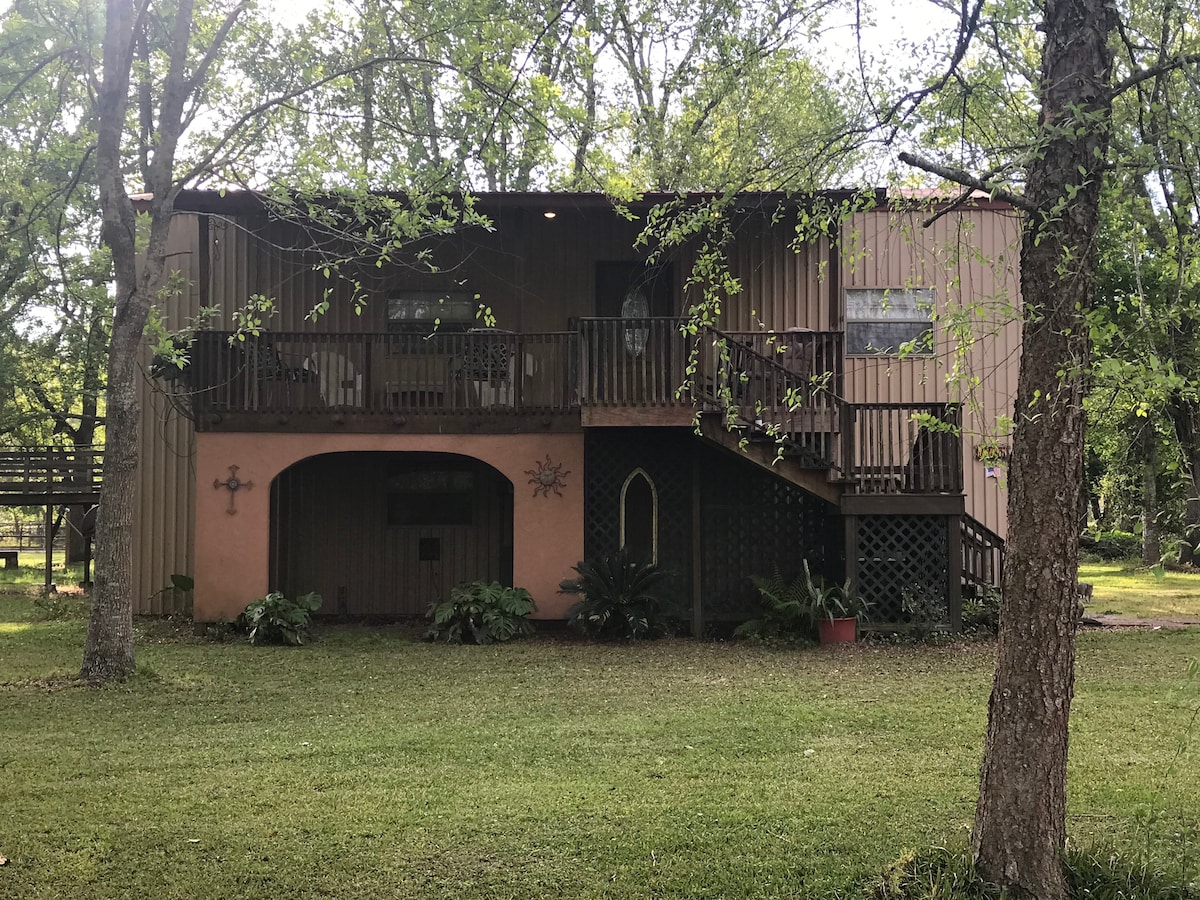 The exterior of a two-story home is surrounded by greenery, featuring a wooden deck and railing. Lush plants are positioned around the base, while large trees provide shade. A decorative sun ornament and a small archway add character to the entrance.