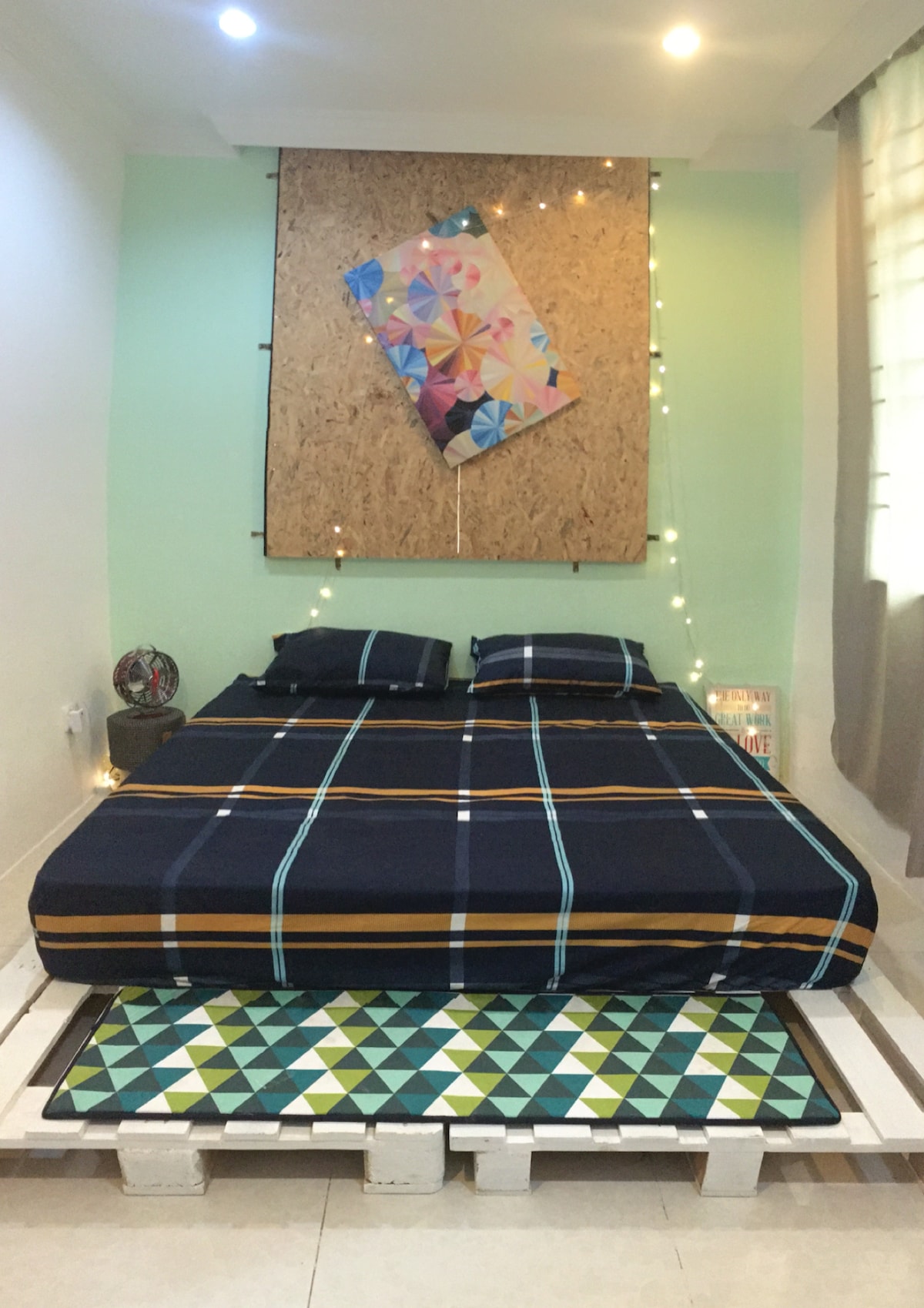 A king-sized bed adorned with a plaid patterned bedspread is positioned against a light green wall, complemented by a colorful piece of wall art. Soft lighting is provided by nearby fairy lights, creating a cozy atmosphere. A patterned mat lies in front of the bed.