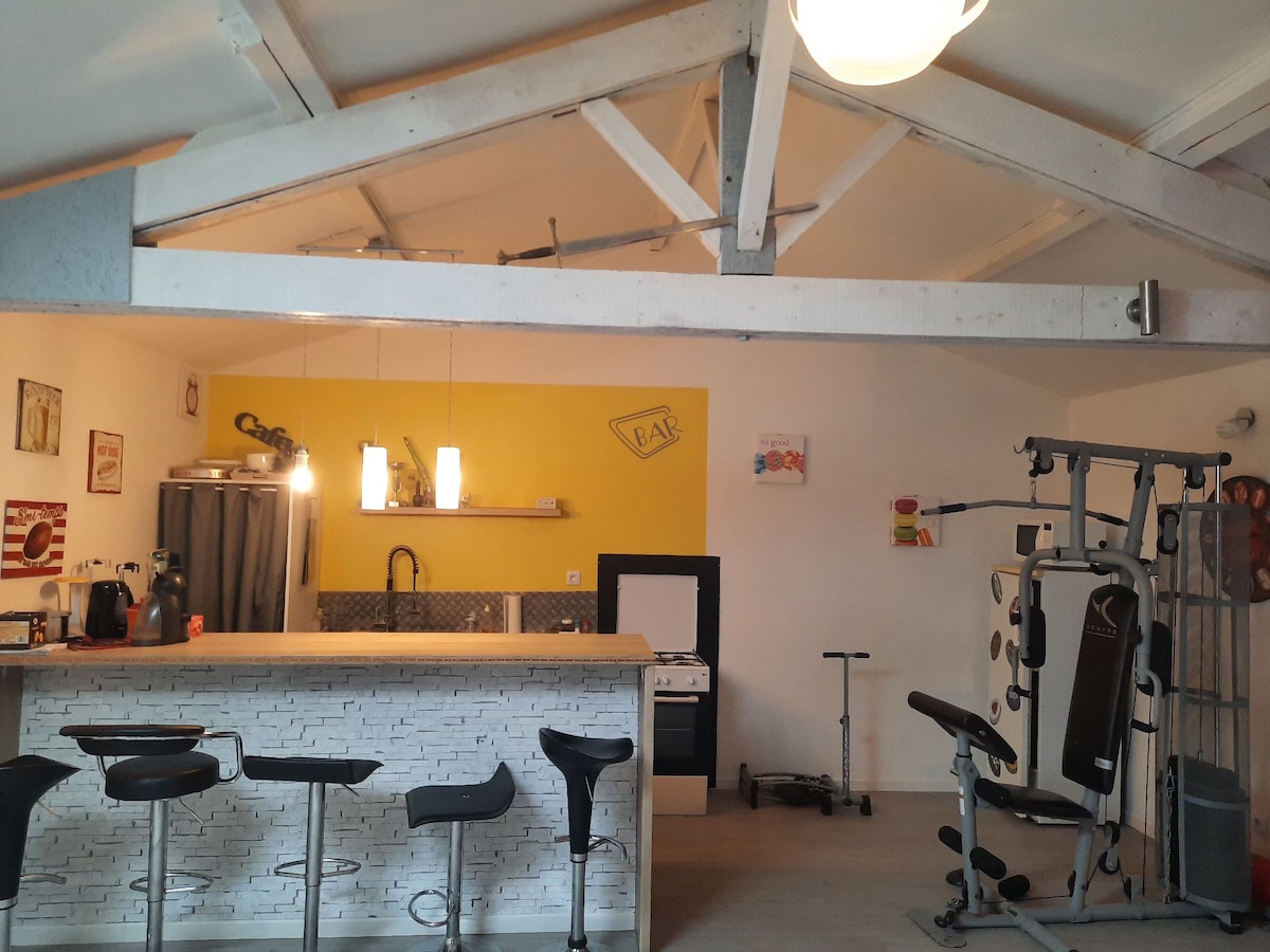 An open-plan kitchenette and living area is displayed, featuring a wooden bar with high stools. A workout station is visible in the corner, with sleek design elements complementing the bright yellow accent wall. Natural light is diffused through a central ceiling fixture.