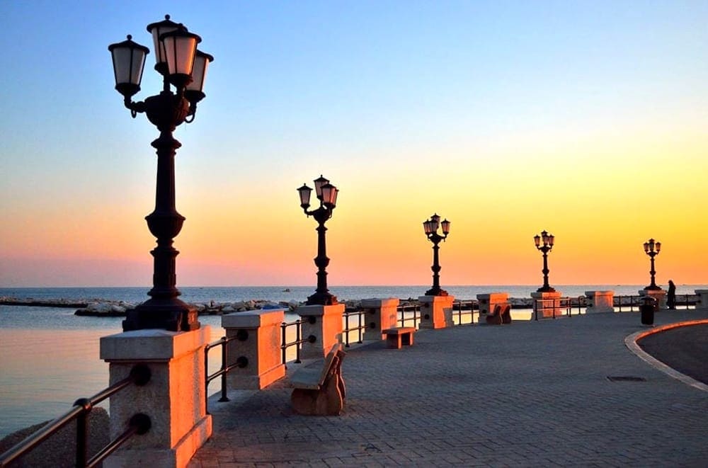 The image captures a tranquil waterfront promenade lined with vintage-style lamp posts. The sky transitions to warm hues of orange and pink, reflecting softly on the calm water. The paved walkway invites leisurely strolls while the distant horizon meets the sea.
