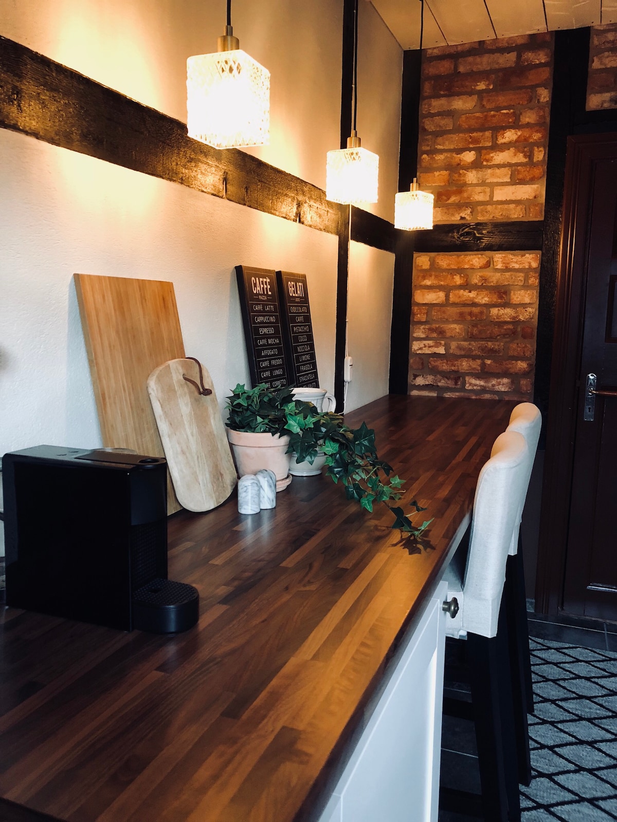 A wooden countertop extends along the wall, complemented by soft pendant lighting above. A coffee maker and cutting boards are positioned neatly, while decorative plants add a touch of greenery. The textured brick wall provides a rustic backdrop to the inviting kitchen space.