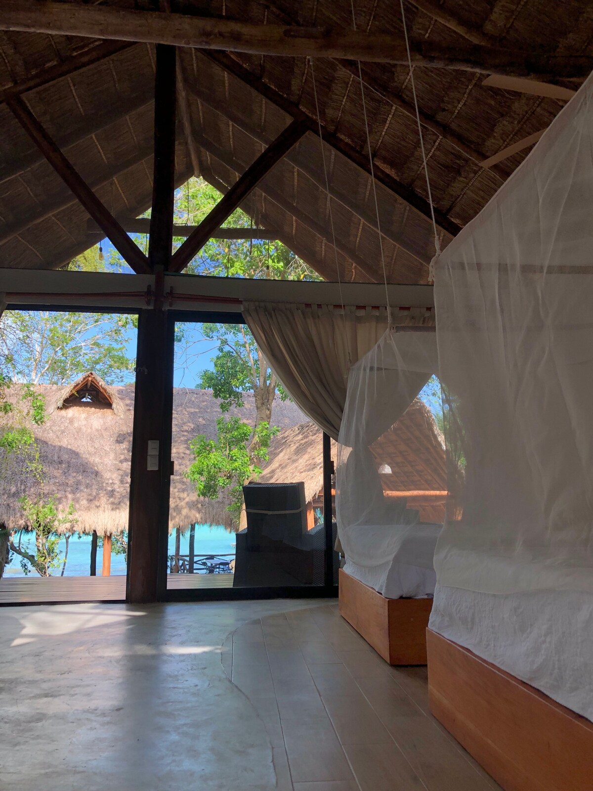 Large windows provide natural light and a view of the surrounding greenery and lagoon. The room features a high thatched roof and wooden accents, creating an open and airy atmosphere. A sheer curtain gently divides the space, offering a touch of privacy.