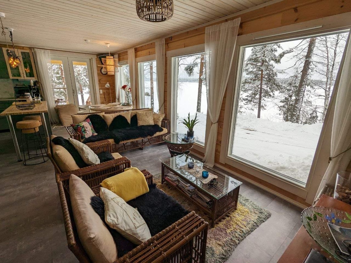 A welcoming living area features comfortable seating made of natural materials, positioned near large windows. Snowy landscapes outside are visible through the expansive glass, filling the space with natural light. Decorative elements include plants and a coffee table adorned with various accents.