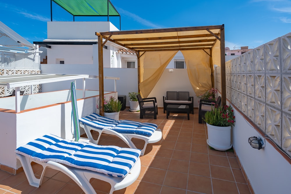 A spacious terrace is shown featuring a pergola and two lounge chairs with striped cushions. An outdoor seating area is present beneath the pergola, surrounded by potted plants. Sunlight brightens the setting, enhancing the inviting feel of the space.
